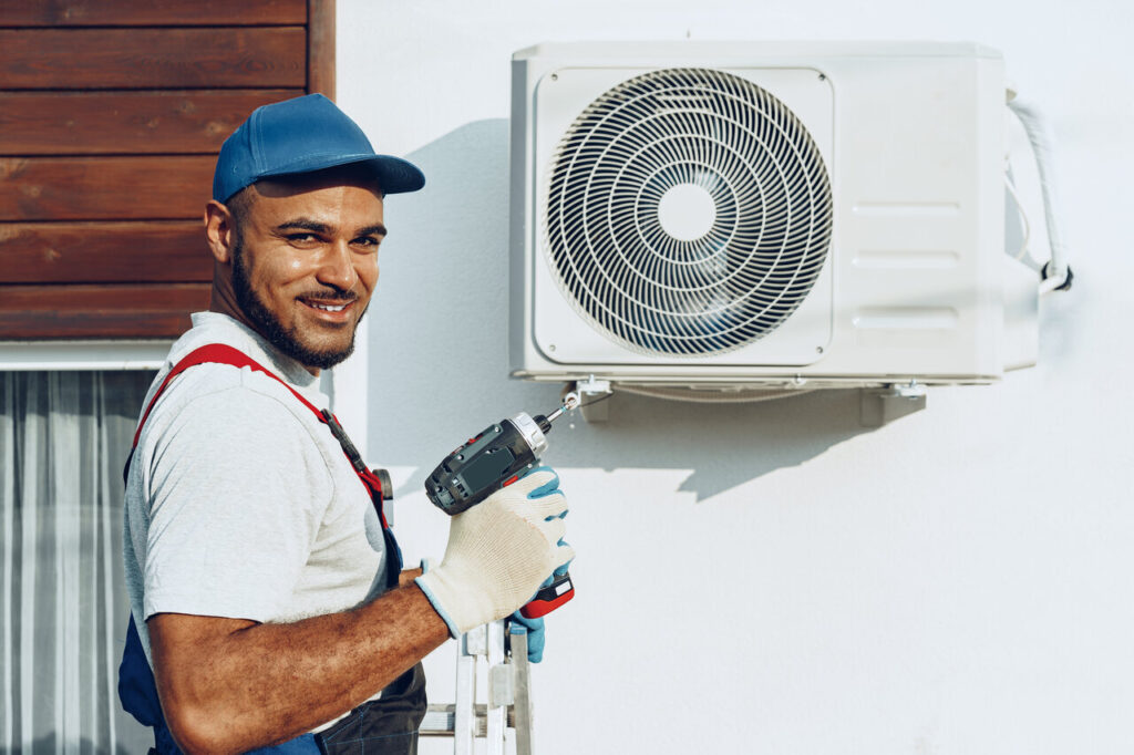 HVAC Installation In Andersonville, TN 1 A man in overalls and a hat holds a tool beside a white air conditioner.