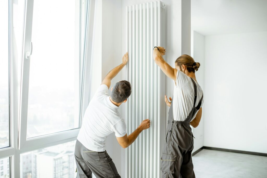 Two men repairing a radiator in an empty room.